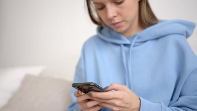 Blonde woman with cellphone focused in hands typing, wearing blue hoodie. Busy freelancer texting in mobile phone working at home. Concept of remote work