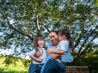 Two sibling little girls with mom laughing and hugging each other on warm and sunny summer day in the garden. Young girls with her mother spending day in park.