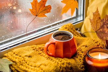 September. Outside the window, autumn leaves and raindrops. Autumn still life with a beautiful bokeh. A cup of hot tea, a candle and a knitted blanket on the windowsill.