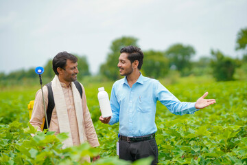 Young indian agronomist giving liquid fertilizer bottle to farmer and saying product information at green agriculture field.