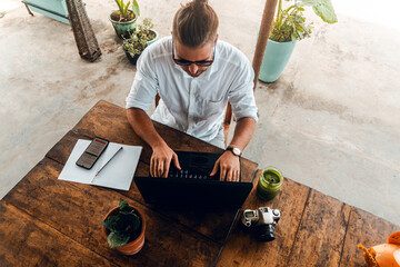 Mężczyzna, cyfrowy nomada pracujący z laptopem i telefonem przy stoliku w kawiarni, praca zdalna podczas podróży. - obrazy, fototapety, plakaty