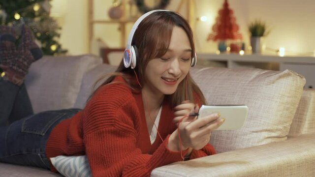 happy woman lying prone on living room sofa with headphones is spending her Christmas binge watching program on phone with a hearty laugh.