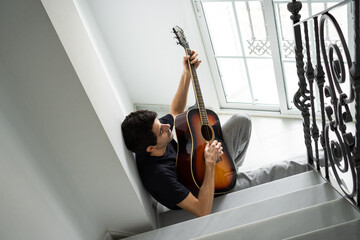 Relaxed young man playing guitar on staircase near window at home.