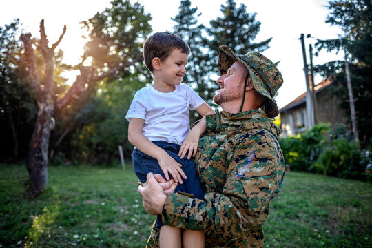 Cropped Shot Of A Young Soldier Men And Son In A Park. Military Man Father Hugs Son. Cute Caucasian Boy Hugs His Dad In The Front Yard. His Dad Has Return From Overseas Military Assignment.