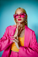 Fashion portrait of beautiful blonde woman wearing trendy pink sunglasses, fuchsia color blazer, orange top, big eaarings, posing on blue backgroumd. 