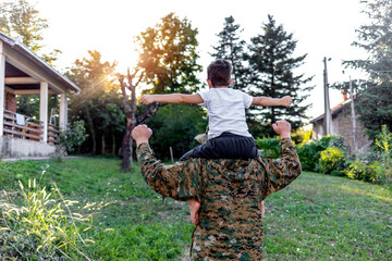 Cheerful dad is glad to be home from military assignment, he is carrying his son on his shoulders. Young military men holding his son on the shoulders with arms raised up in the air, looking away.