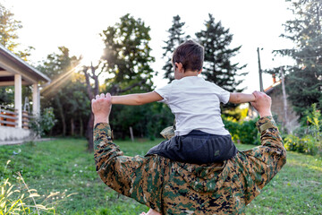 Cheerful dad is glad to be home from military assignment, he is carrying his son on his shoulders. Young military men holding his son on the shoulders with arms raised up in the air, looking away.