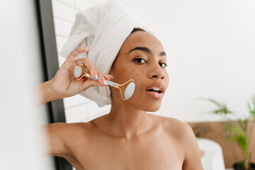 Young black woman wrapped in towels massaging face with jade roller