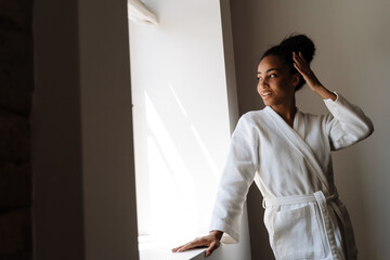 Young black woman wearing bathrobe smiling while posing by window - obrazy, fototapety, plakaty