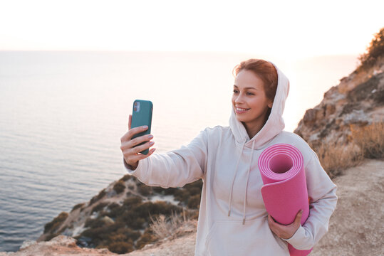 Smiling adult girl 24-26 year old training wear beige hoodie sweatshirt holding yoga mat make photo with phone over sea nature background. Healthy lifestyle. 20s. Happy woman doing fitness on rock.