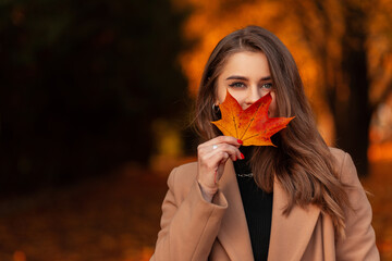 Happy beautiful woman in a fashionable beige coat and sweater covers her face with a colored autumn...