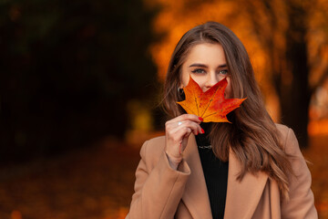 Happy beautiful woman in a fashionable beige coat and sweater covers her face with a colored autumn leaf in a park with orange fall foliage. Space for text
