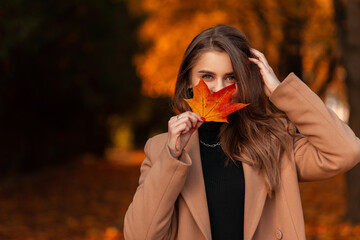 Happy beautiful young girl in fashionable coat covers her face with colored autumn leaf in the golden fall park. Autumn holidays