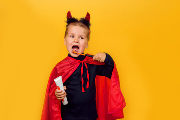 One little toddler boy in a carnival costume with toothpaste and an orange brush for Halloween is isolated on a yellow background. Medicine, dental hygiene, holidays concept.