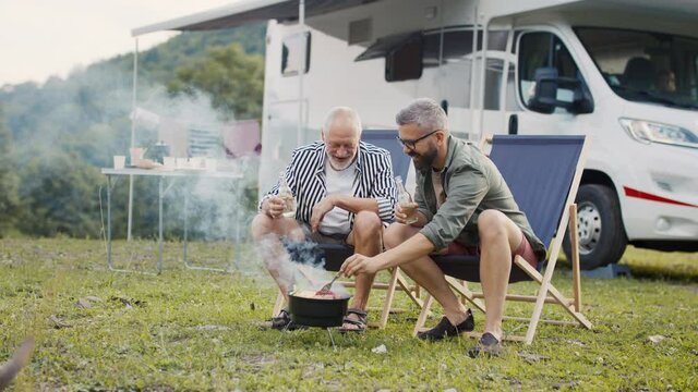 Mature man with senior father talking at campsite outdoors, barbecue on caravan holiday trip.