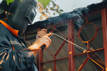 Close up of a welder hands welding steel structure metal part in home backyard. Craft man use electric arc argon wear work uniform,protective helmet on head without gloves while sparks fly at factory.