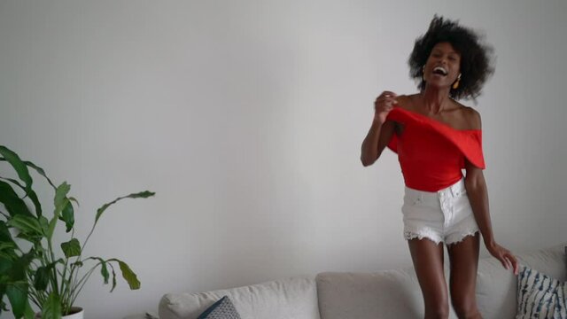 Happy Black woman with curly hair having fun jumping dancing on the couch at home