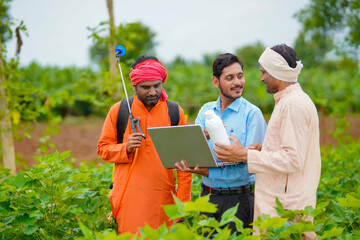 Young indian agronomist giving liquid fertilizer bottle to farmer and showing product information in laptop at green agriculture field.