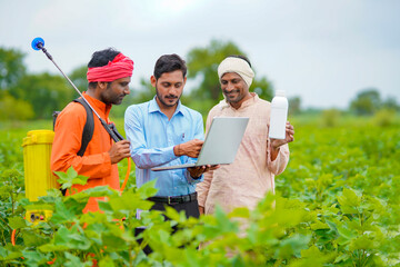 Young indian agronomist giving liquid fertilizer bottle to farmer and showing product information in laptop at green agriculture field.