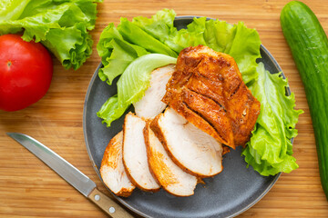 Grilled poultry fillets are sliced on a plate with lettuce. Nearby vegetables: cucumber and tomato.