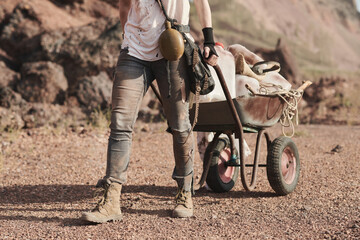 Close-up of female refugee in dirty clothing carrying wheelbarrow with things behind her in the desert