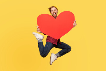 happy in love man jumping and holding big red heart shape and looking with love and screaming. indoor studio shot isolated on yellow background