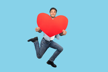 happy in love man jumping and holding big red heart shape and looking with love and toothy smile. indoor studio shot isolated on blue background