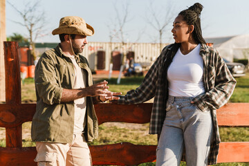 Multiracial man and woman talking while standing by fence on farm