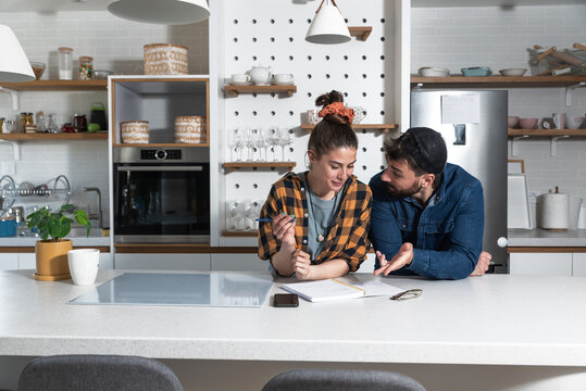 Young Happy Couple Bride And Groom Standing In Their Kitchen And Want To Make A List Of People, Family And Friends Who They Will Invite To Their Wedding Ceremony