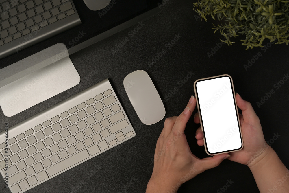 Poster closed up with hand holding a white blank screen smartphone over a working desk surrounded by variou