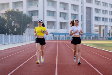 Obraz premium Two young Asian women in sports outfits jogging on running track in city stadium in the sunny morning for a healthy lifestyle. Young fitness women run on the stadium track.