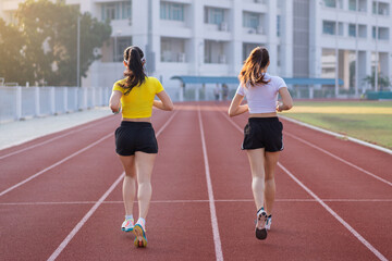 Obraz premium Two young Asian women in sports outfits jogging on running track in city stadium in the sunny morning for a healthy lifestyle. Young fitness women run on the stadium track.