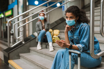 Multiracial two women in face masks sitting on stairs at train station