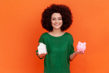 Smiling woman with Afro hairstyle wearing green casual style sweater standing holding piggy bank...
