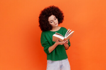 Attractive concentrated woman with Afro hairstyle wearing green casual style sweater reading interesting book, being very attentive. Indoor studio shot isolated on orange background.