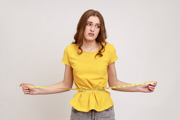 Portrait unhappy smiling woman in yellow T-shirt measuring her waist with tape measure and expressing sadness, looking away with frowning face. Indoor studio shot isolated on gray background.