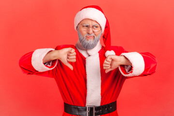 Fototapeta na wymiar Elderly man with gray beard wearing santa claus costume has naughty expression and showing thumbs down, expressing disapproval, negative feedback. Indoor studio shot isolated on red background.