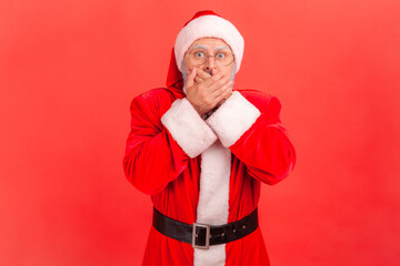 Portrait of elderly man with gray beard wearing santa claus costume closing mouth with his hands,...