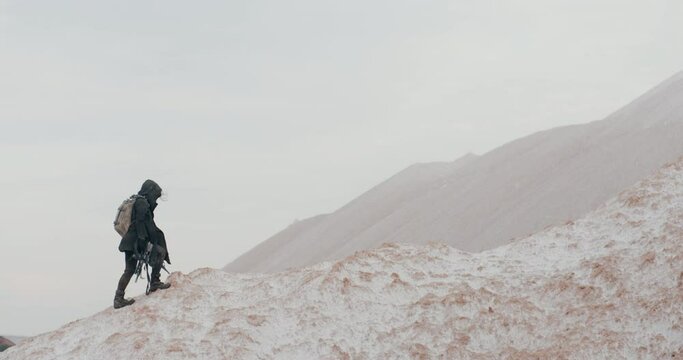 Wide shot post apocalyptic woman in grunge clothes hooded black mantle protective gas mask respiratory walking up hill in white desert. Cyberpunk soldier holding assault gun. Future female survivor