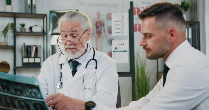Handsome positive confident 40-aged bearded man visitingmedical office where responsible experienced senior grey-haired doctor discussing with him results of x-ray scan