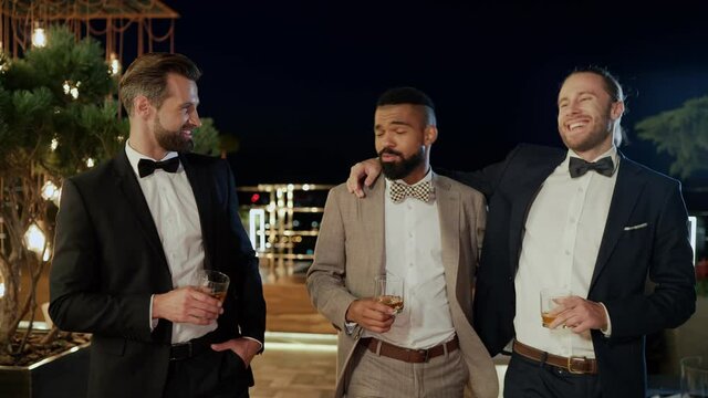 Three happy men in suits holding glasses with while spending time on party