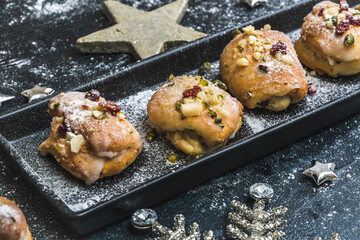 Homemade mini stollen with icing, dried cranberries and pistachios on a black tray on a black background, decorated with silver stars.