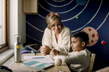Smiling mother looking at autistic son sitting at table in home