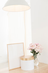 Vertical frame and gift box mockup on a wooden table in the kitchen. Glass jug with a bouquet of pink peonies. Scandinavian style interior.