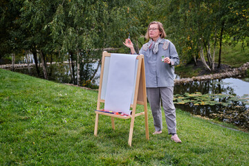 Happy woman draws on the easel with a brush and paints. Woman artist draws nature and trees on paper by the water on the river bank