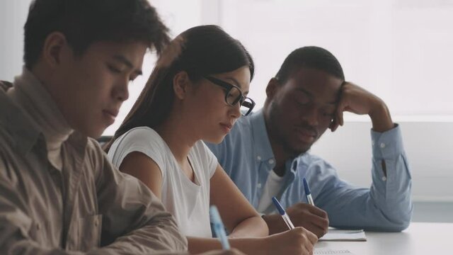 University life. Focused diverse students writing in copybooks, sitting in row at college classroom, slow motion