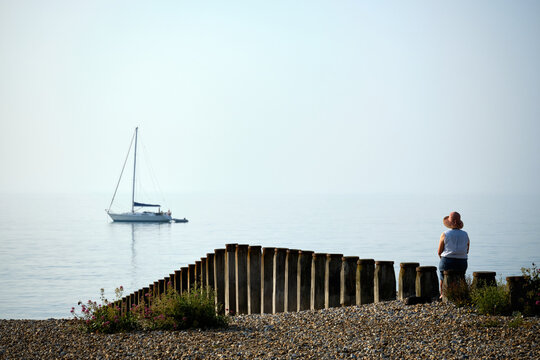 Eastbourne, East Sussex, United Kingdom - July 20 2021:  High-key image of woman looking at a yacht in calm sea off Eastbourne beach in East Sussex as the morning mist lifts.