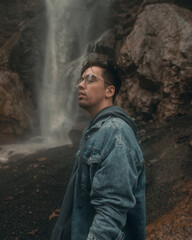 young guy in a denim jacket against the backdrop of a waterfall
