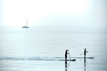 Two paddle boarders on a calm early morning sea in Eastbourne with a yacht in the distance.