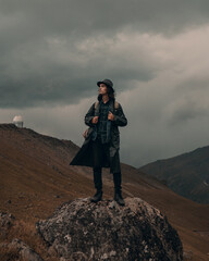 young guy in a raincoat and hat stands on a stone in the mountains
